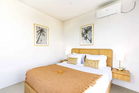 Cozy tropical bedroom with wooden bed, white linens, orange fringed throw, bedside lamps, and black-and-white palm tree artwork on white walls.
